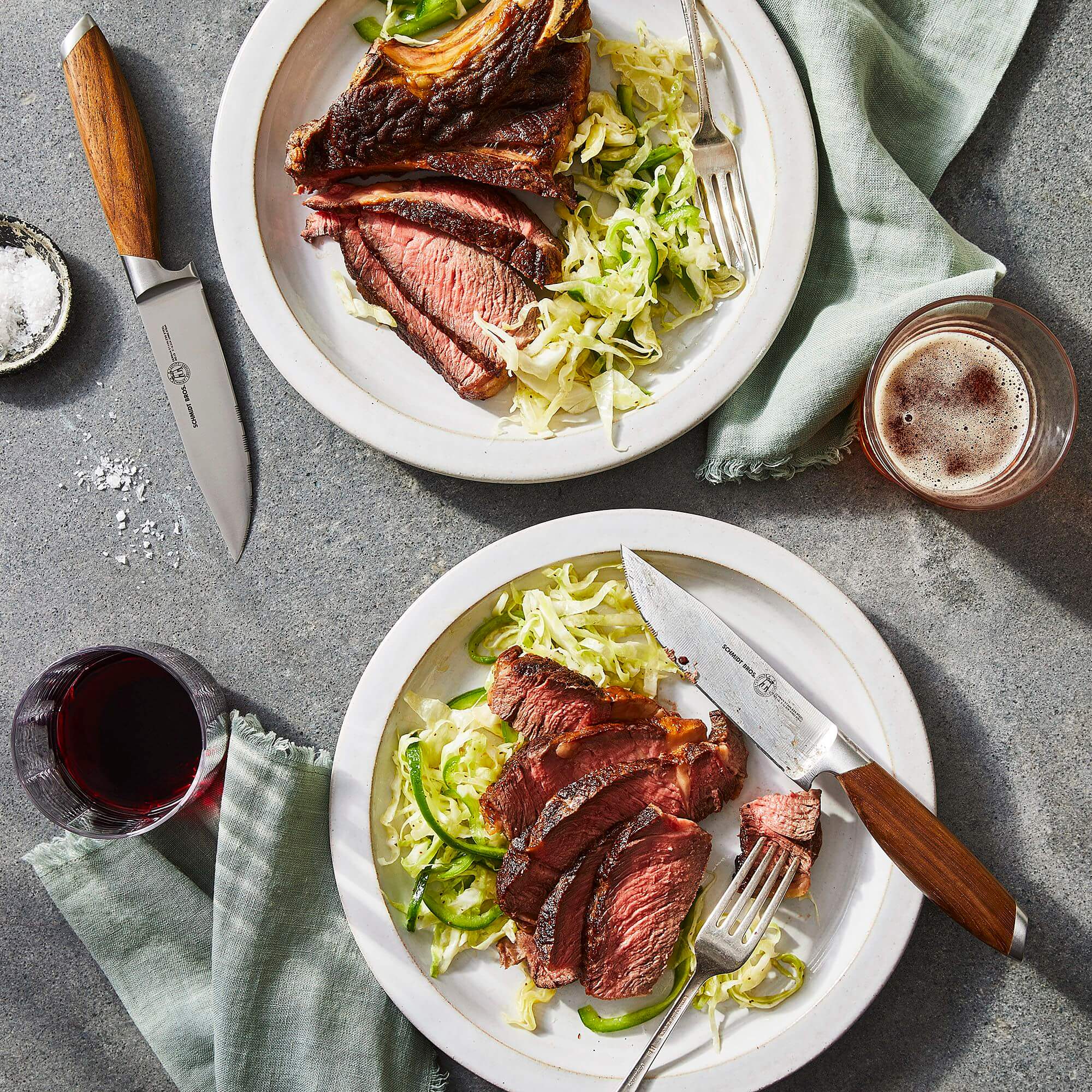 Two plates of grilled steak with carrot top chimichurri sauce served with shredded greens and accompanied by knives and drinks.