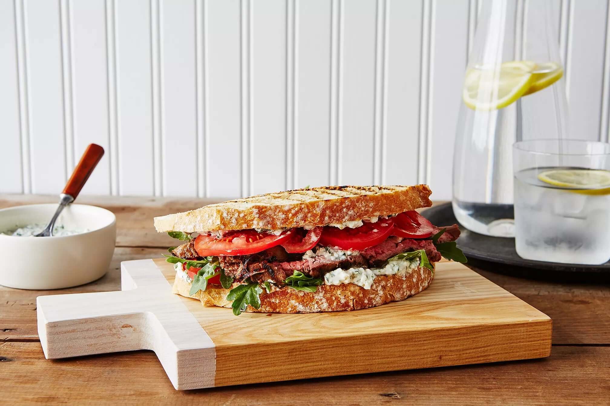 Best steak and feta sandwich on wooden board, garnished with tomatoes and greens, with a glass of lemon water in the background.